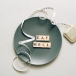Flat lay of scrabble tiles spelling 'eat well' on a plate with a tape measure, promoting healthy eating.