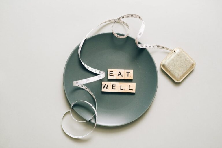 Flat lay of scrabble tiles spelling 'eat well' on a plate with a tape measure, promoting healthy eating.