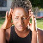A woman holding her head in pain, indicating a severe headache outdoors in a sunny setting.